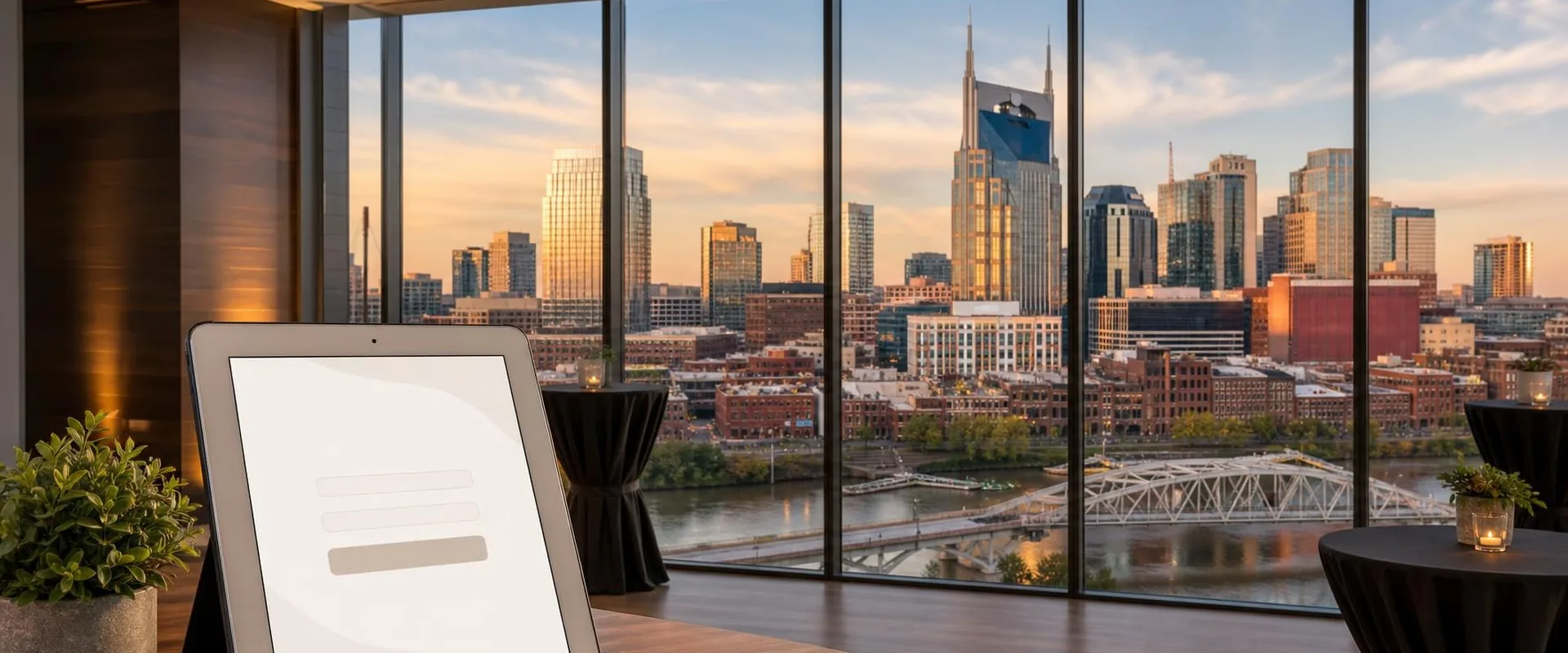 iPad check-in screen in a black folio stand on a table in a high-rise Nashville event space, with the AT&T Building, the downtown skyline and the Cumberland River behind it